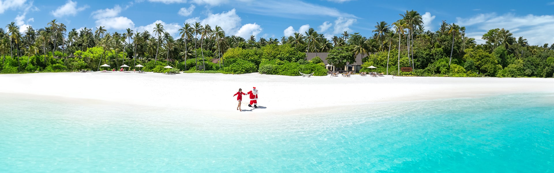Santa walking on the sandy white beach with beach pavilions at the backdrop at Avani+ Fares Maldives Resort